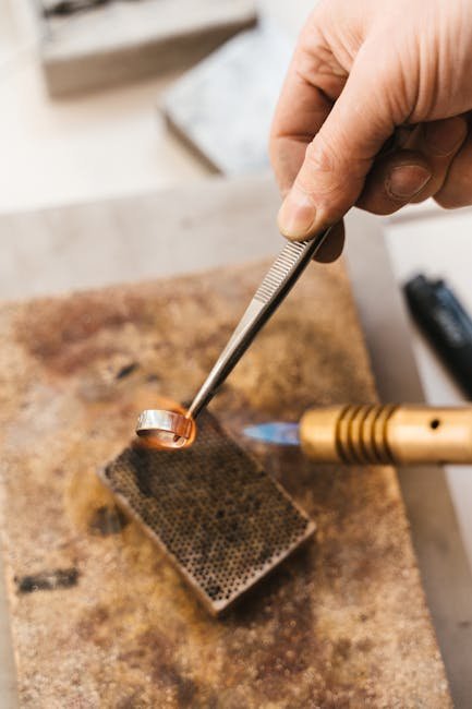 A jeweler uses a blowtorch and tweezer to craft a metal ring in a workshop setting.